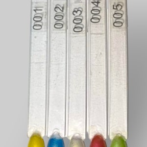 Puede incluir: Cinco muestras de esmalte de uñas en palitos blancos. Las muestras están etiquetadas como 001, 002, 003, 004 y 005. Los colores son amarillo, azul, blanco, rojo y verde.
