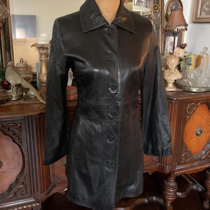 Puede incluir: Una taza de cerámica blanca con un asa negra y el texto "But First, Coffee" en negro, con una fuente de estilo manuscrito. La taza está sobre una superficie blanca.