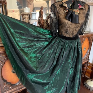 以下が含まれることがあります： ヴィンテージのエメラルドグリーンのスカートとトップのアンサンブル。スカートは流れるような玉虫色の生地で、トップスは黒いレースの袖と黒いベルベットのウエストバンドが特徴です。首には花の模様のスカーフが巻かれています。