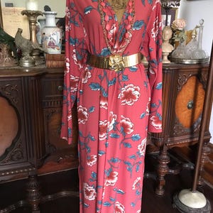 May include: A coral-colored dress with a floral pattern of red and white flowers and blue leaves. The dress has long sleeves, a V-neck, and a gold belt. A gold necklace and pink beaded necklace are also visible.
