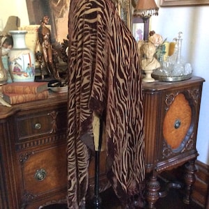 May include: A brown and tan zebra-striped velvet poncho featuring a textured, burnout-style pattern with a soft, draped silhouette and fringe detailing along the bottom hem, displayed on a mannequin stand.