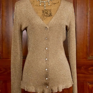 Puede incluir: Un cárdigan dorado de manga larga con cuello en V y cierre de botones. El cárdigan tiene un brillo sutil y un dobladillo con volantes. Un delicado collar se muestra en un maniquí. El fondo es una puerta de madera oscura.