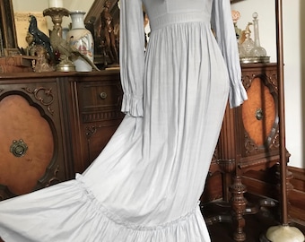 Könnte beinhalten: Ein hellblaues langärmeliges Maxikleid mit hochgeschlossenem Knopfkragen, Rüschendetails an den Schultern und einer gerafften Taille. Der Rock hat einen breiten Rüschensaum am unteren Ende und die Ärmel enden mit gerüschten Manschetten.