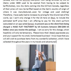 May include: A person with long hair wearing a light blue long-sleeved shirt and blue jeans kneeling on a porch in front of a large cardboard box. The person has both hands raised in the air with palms facing forward near a dark grey front door.