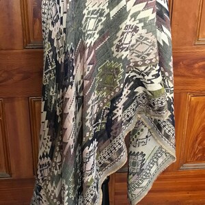 Può includere: Sciarpa fantasia con un design geometrico nei toni del grigio, verde e beige. La sciarpa è drappeggiata su un manichino nero, mettendo in mostra i suoi bordi sfrangiati e il suo motivo intricato. Il design della sciarpa include forme a diamante e a zig-zag.