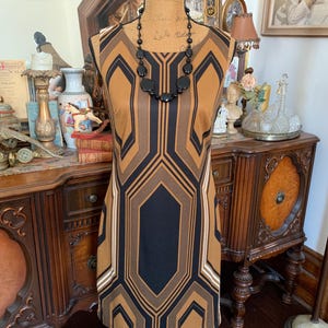 May include: A sleeveless dress with a geometric pattern in brown, black, and tan. The dress features a black central panel and white stripes. A black beaded necklace is worn. The dress is displayed on a mannequin.