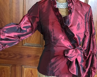 May include: A burgundy wrap blouse with long, gathered sleeves and a high ruffled collar. The blouse features a bow at the waist and a shimmering, textured fabric. A silver necklace is visible at the neckline.