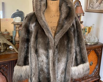 May include: A brown and grey faux fur jacket featuring a large shawl collar and long sleeves with contrasting light-coloured fur cuffs. The jacket has a thick, textured pile and is displayed on a mannequin torso, showcasing the open front design.