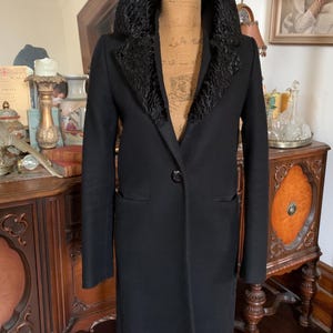May include: A black coat featuring a prominent, textured black faux fur collar. The coat has long sleeves, a single central button closure, and two side pockets. It is displayed on a beige mannequin torso, showcasing a structured silhouette.