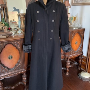 May include: A long black coat featuring a high collar with dark grey faux fur lining, matching faux fur cuffs, and a double-breasted front closure with six round metallic buttons. The coat has a structured fit and reaches below the knees.