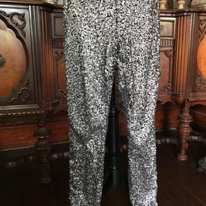 May include: Full-length, silver sequin trousers. The trousers are covered in small, reflective sequins that create a shimmering effect. The trousers are displayed on a black stand, showcasing their straight-leg design. The background features antique furniture.