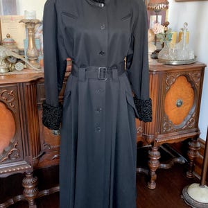 May include: A long, black coat with a high collar and a belted waist. The coat has a textured collar and cuffs, and a row of buttons down the front. The coat is a classic style, suitable for formal occasions.