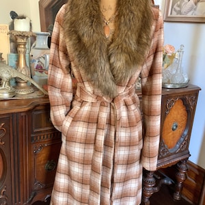 May include: A long, plaid coat in shades of brown and cream, with a faux fur collar and a matching belt. The coat is displayed on a mannequin, showcasing its full length and design. The coat has pockets and a tie belt.