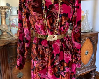 May include: A long-sleeve velvet wrap dress featuring a vibrant abstract pattern in shades of magenta, deep red, and black. The dress is cinched at the waist with a gold-toned metal mesh belt and accessorised with a long beaded gold chain necklace.