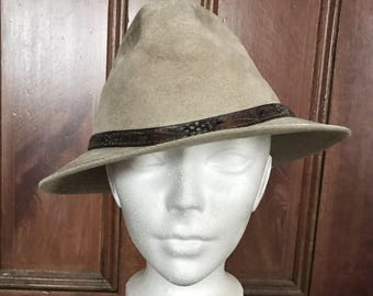 May include: A tan fedora hat with a brown band featuring a feather design. The hat has a classic, slightly worn appearance, with a soft texture. The brim is moderately sized, and the crown is indented.