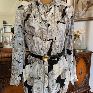 Könnte beinhalten: Langärmliges Hemd mit floralem und musikalischem Druck in Schwarz-, Weiß- und Grautönen. Das Hemd wird mit einem schwarzen Gürtel mit goldfarbener Schnalle in der Taille zusammengehalten. Das Hemd hat einen hohen Kragen.