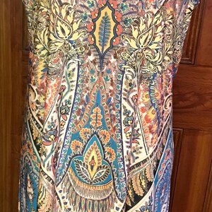 Peut inclure: Une robe sans manches avec un motif paisley bleu, orange et blanc. La robe a une ourlet inférieur bleu turquoise et un collier bleu turquoise avec de grandes pierres ovales.