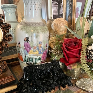 Peut inclure: Un vase en porcelaine blanc avec une peinture détaillée de personnages en costume traditionnel, sur fond de bambous. Une pochette noire à perles est devant le vase, avec une rose rouge et d'autres éléments décoratifs.