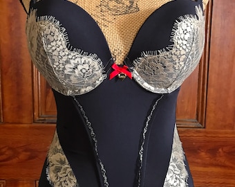 May include: Navy blue bustier with beige lace detailing on the cups and sides. A small red bow accents the center. The bustier is displayed on a mannequin with a silver necklace.