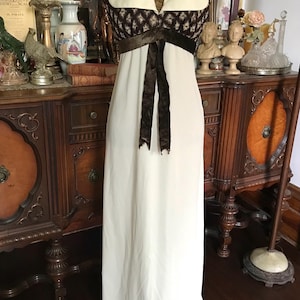 Puede incluir: Vestido vintage color marfil con cuello halter. El corpiño presenta un diseño marrón y crema estampado, acentuado con una cinta de terciopelo marrón oscuro. El vestido se exhibe en un maniquí en una habitación con muebles antiguos.
