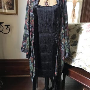 Puede incluir: Un vestido negro con flecos se exhibe en un maniquí. Un kimono con estampado floral en tonos verdes, rojos y negros está drapeado sobre el vestido. Se ve un collar con un colgante.