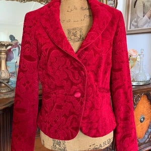 Peut inclure: Un blazer rouge avec un motif floral. Le blazer a une fermeture à un seul bouton et un revers cranté. Le tissu a un aspect texturé, semblable à du velours. Le blazer est présenté sur un mannequin.