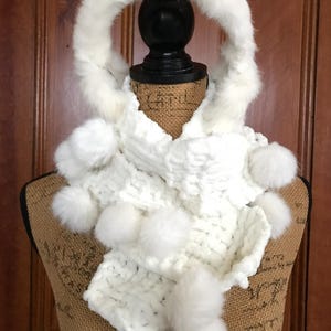 May include: A white knitted scarf with a hood and fluffy pom-poms. The scarf is displayed on a mannequin, showcasing its texture and design. The hood is trimmed with a soft, white fur.