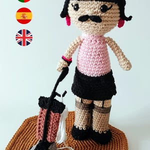 May include: A handmade crochet doll with a black wig, mustache, and pink earrings. The doll wears a pink top, black skirt, and patterned stockings. It holds a miniature vacuum cleaner and stands on a brown crocheted mat.