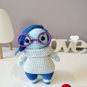 May include: A blue and grey crocheted stuffed toy with purple glasses. The toy is wearing a grey sweater and blue pants. The toy is standing on a white surface with colorful crocheted balls.
