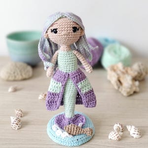 Könnte beinhalten: Eine handgefertigte Häkel-Meerjungfrauenpuppe mit hellviolettem Haar, einem blassgrünen Oberteil und einem violetten Rocküberzug. Die Puppe steht auf einer kreisförmigen hellblauen Basis, die mit einem kleinen braunen Fisch und weißen Wellen verziert ist.