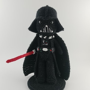 Puede incluir: Figura de Darth Vader negra hecha a mano con ganchillo y un sable de luz rojo. La figura tiene un casco detallado, capa y peto. Se apoya sobre una base redonda negra, mostrando un coleccionable único de Star Wars hecho a mano.