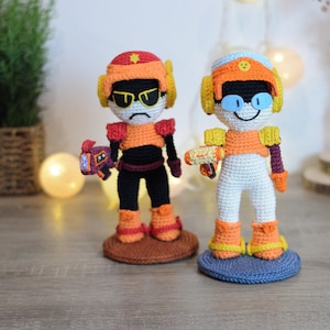 Op de afbeelding: Twee handgemaakte gehaakte figuren van een robotachtig personage. Eén figuur draagt een zwarte broek en de andere een witte broek. Beide figuren hebben oranje en gele accenten en houden een speelgoedpistool vast.