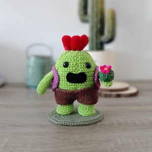Op de afbeelding: Een handgemaakte gehaakte cactusfiguur in groen, bruin, paars en rood. De figuur heeft een lachend gezicht, een rode bloem bovenop en houdt een kleine cactus met een roze bloem vast. Hij staat op een groene gehaakte basis.