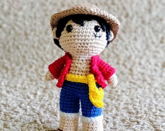 May include: A crocheted doll with brown hair, wearing a red shirt, blue pants, a yellow belt, and a brown straw hat.