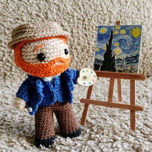 Könnte beinhalten: Eine gehäkelte Figur eines Menschen, der einen braunen Hut, ein blaues Hemd und eine braune Hose trägt, eine Farbpalette hält und vor einem Miniatur-Staffelei mit einem Gemälde von Van Goghs Sternennacht steht.