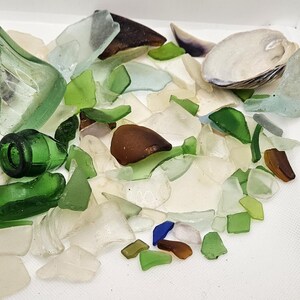 May include: A collection of sea glass in various shapes and sizes, including green, white, and brown pieces. Also includes a seashell and a small, green glass bottle. The sea glass is arranged on a white surface.
