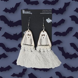May include: White ghost-shaped wooden earrings with white tassel fringe. The earrings are on a black card with the text "Sonoran Charm Jewelry" and "Stainless Steel Custom Acrylic SonoranCharm.com".