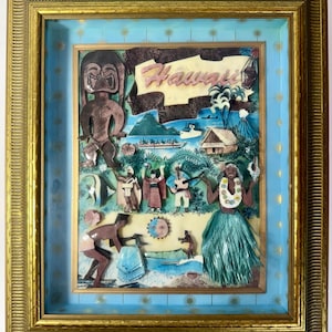 Puede incluir: Una obra de arte en caja de sombra tridimensional enmarcada que presenta un collage en capas de Hawái. La escena incluye una figura tiki, una bailarina de hula con falda verde, músicos, un barco, una choza y un volcán sobre un fondo azul claro.