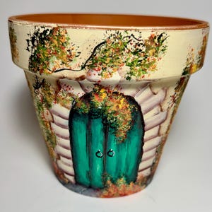 May include: Hand-painted terracotta flower pot featuring a vibrant green door with two silver handles, set against a backdrop of a stone wall and lush foliage. The pot's rim is painted cream, with detailed artwork.