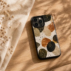Könnte beinhalten: Eine Smartphone-Hülle mit einem Muster aus abgerundeten, kieselsteinartigen Formen in Schwarz, Dunkelgrün, Beige und gebranntem Orange auf weißem Grund mit schwarzen Sprenkeln. Die Hülle hat einen schwarzen Rahmen und eine Aussparung für drei Kameras.
