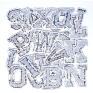 May include: A collection of silver sequin letters, including the letters O, U, E, N, I, T, W, A, K, X, L, and P. The letters are arranged in a random order and are all the same size.