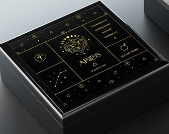 May include: Black wooden box with a gold design featuring the zodiac sign Aries. The box is open and the inside is lined with white felt.