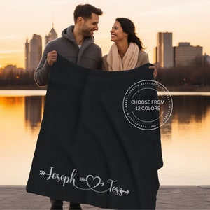 May include: A black fleece blanket with the names "Joseph" and "Tess" written in white cursive with an arrow pointing to each name and a heart connecting the two names. The blanket is held by a couple standing in front of a lake and city skyline. The text "Choose from 12 colors" is printed on a white circle on the blanket.