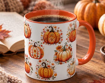May include: A white ceramic mug with an orange handle and rim, featuring a repeating pattern of orange pumpkins and autumn leaves. The mug is filled with a dark liquid and sits on a wooden surface, showcasing a vibrant seasonal design.