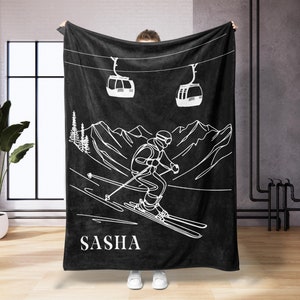 Könnte beinhalten: Schwarze Fleece-Decke mit einer weißen Umrandung eines Skifahrers auf einer Bergabfahrt, zwei Skiliften und dem Namen "SASHA" unten.