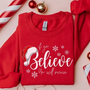 May include: A red sweatshirt featuring the words "If you Believe You will receive" and a Santa hat illustration. Christmas decorations, such as baubles and a wrapped present, surround the garment.