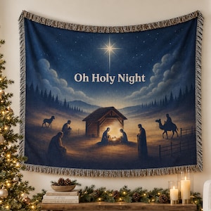 May include: A woven tapestry throw blanket featuring a dark blue night sky with a bright star above a nativity scene. The text Oh Holy Night is printed in white in the centre. The blanket has a fringed edge and depicts figures, a stable, and a camel.