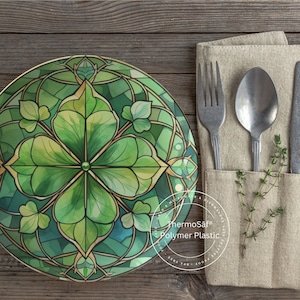 May include: A decorative plate with a stained-glass clover design in shades of green and gold. The plate is next to a linen napkin with a fork, spoon, and knife. A sprig of herbs adds a natural touch to the table setting.
