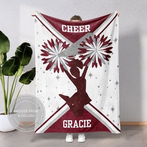 May include: A white fleece blanket with a maroon and gray cheerleading design. The blanket features a silhouette of a cheerleader holding pom-poms, stars, and the words "CHEER" and "GRACIE".