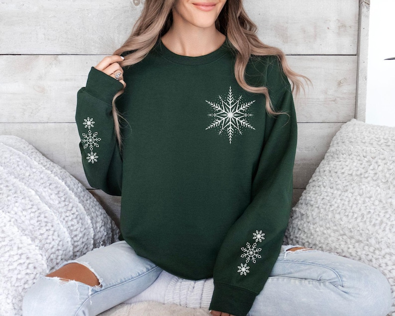 May include: A dark green sweatshirt with white snowflake designs on the sleeves and chest. The sweatshirt is made of a soft, comfortable material and has a relaxed fit.